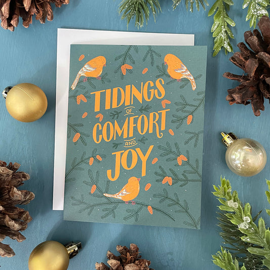 A greeting card features yellow hand-lettering reading tidings of comfort and joy on a blue background. There are painted goldfinches and pinecone-laden branches around the words. The card lies against a white envelope and is surrounded by gold ornaments and pinecones.