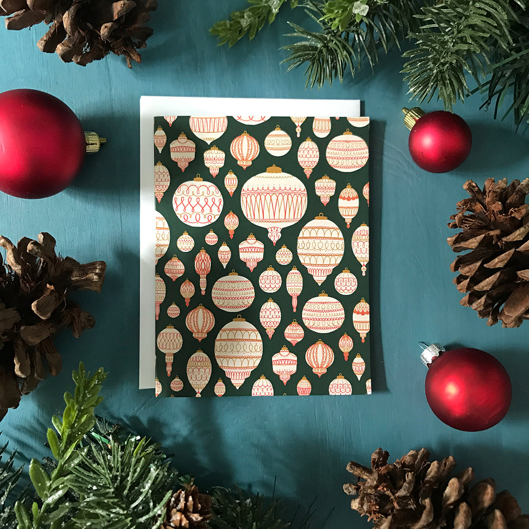 A green greeting card boasts a pattern of cream, red and gold tree ornaments. The card is surrounded by red Christmas ornaments, pinecones, and faux winter greenery on a blue wooden background.