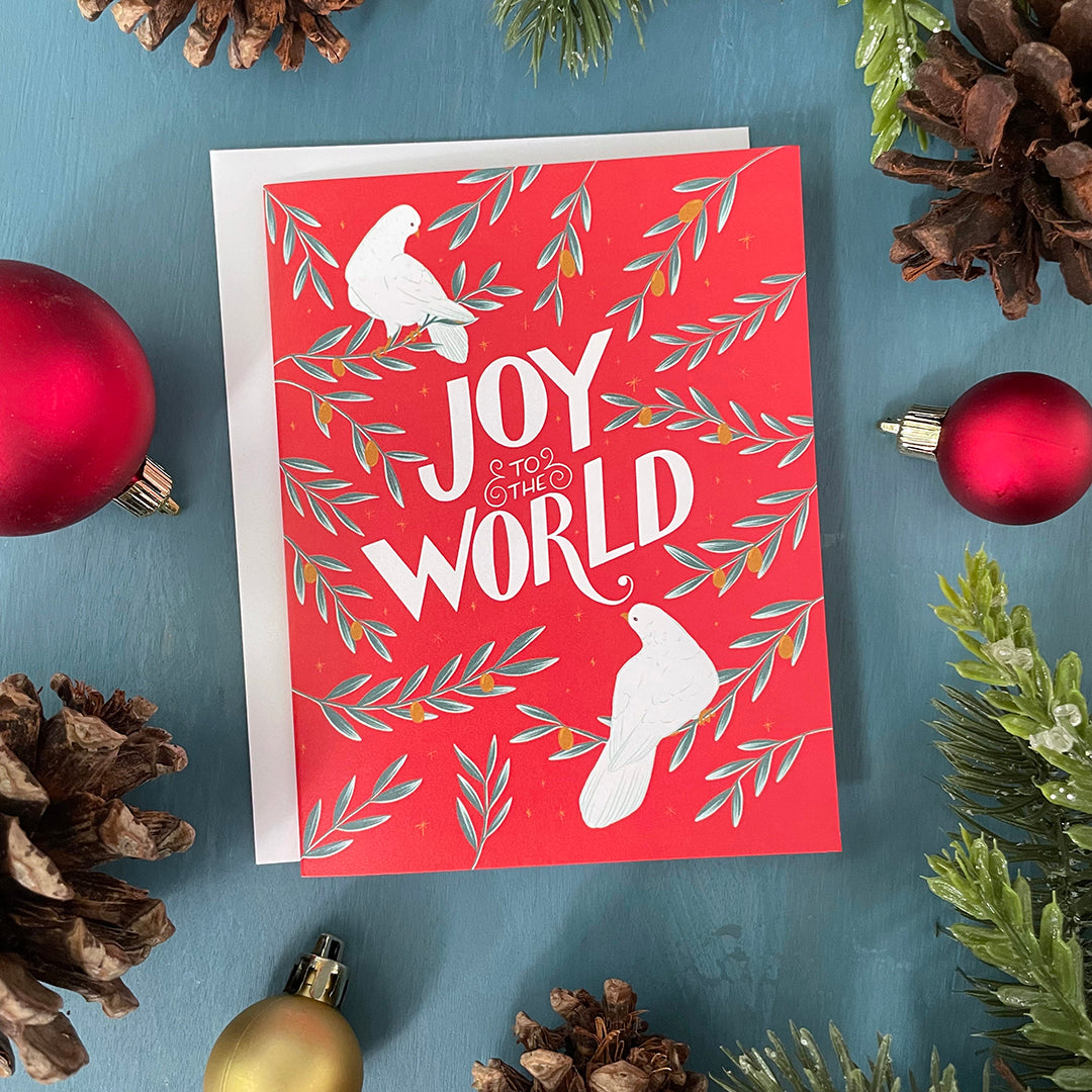 A red greeting card shows two white doves, branches of golden olives, and the hand-lettered words Joy to the World in white. The card is surrounded by ornaments, pinecones, and faux greenery.