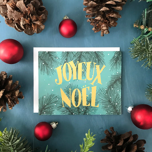 A teal greeting card features the words Joyeux Noel in midcentury-inspired lettering against a background of snowy evergreen branches. The card is surrounded by red Christmas ornaments, pinecones, and faux greenery on a blue background.
