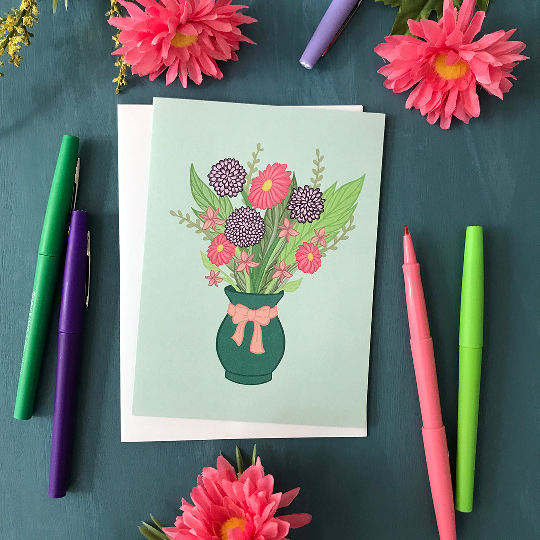 A pastel mint greeting card features an illustration of a bouquet in a green vase with a pink bow around it. The bouquet includes purple and pink flowers and greenery in various shades. The card sits among felt-tip pens in various colours and bright pink artificial flowers.