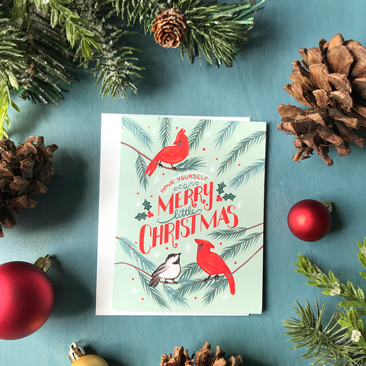 A greeting card reads Have Yourself a Merry Little Christmas lettered in red and green, surrounded by cardinals and a chickadee on a mint background. The card is surrounded by pinecones, Christmas ornaments and faux evergreen branches.