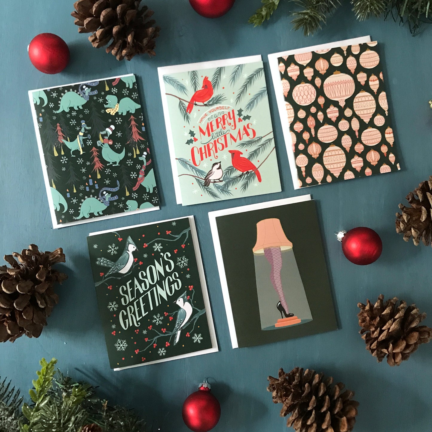 5 Christmas cards - one with dinosaurs, one with cardinals, one with ornaments, one with blue jays, and one with a leg lamp - are laid out among ornaments and greenery on a blue surface.