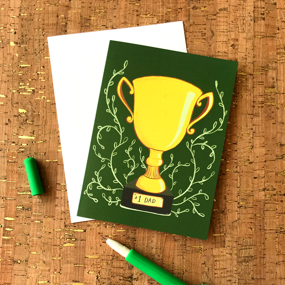 A green greeting card features a yellow trophy with vines coming out of it and number one dad at the bottom. It is against a white envelope on a cork background.