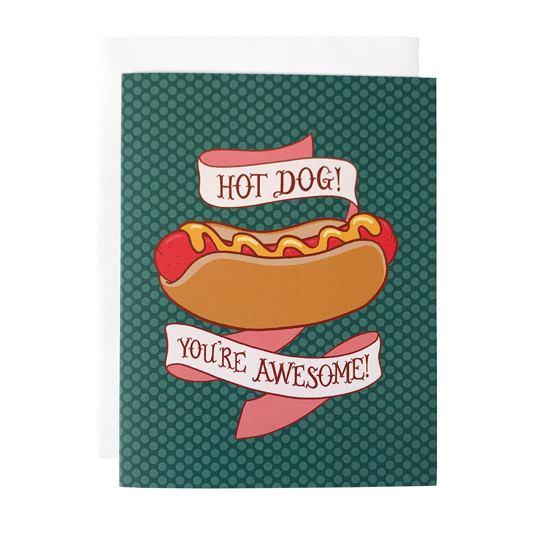 A deep blue-green greeting card shows an ilustration of a hot dog slathered with mustard, surrounded by a banner that reads Hot dog! You're awesome! in capital letters.The card sits against a white envelope on a white background.