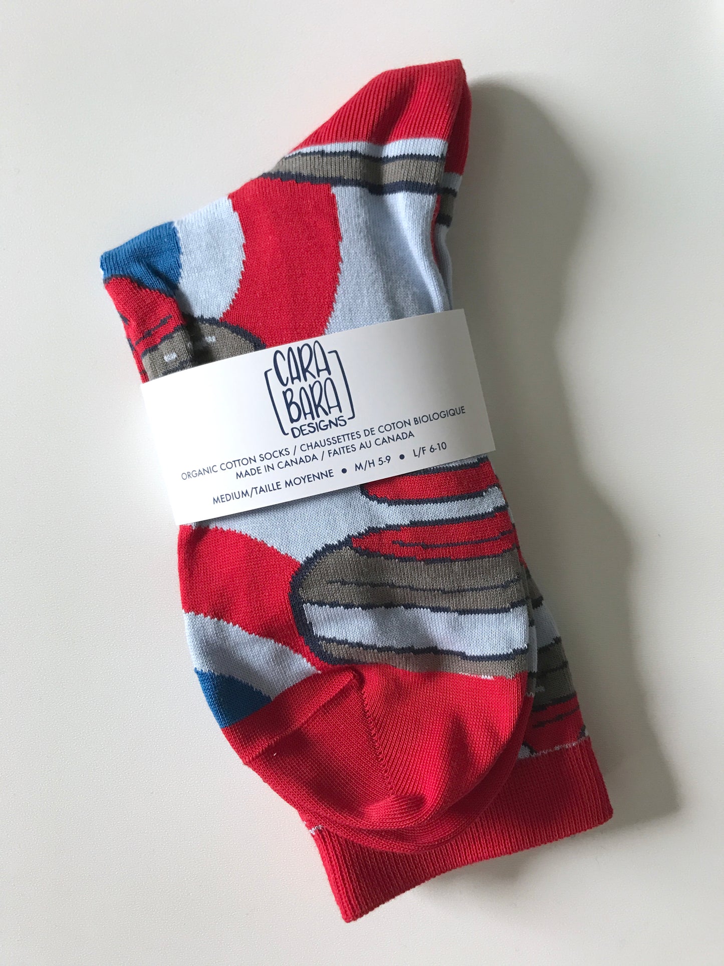 A pair of red and blue socks patterned with curling rocks is folded and labelled with a belly band with the Carabara Designs logo and the words organic cotton socks made in Canada, size medium, in English and French.