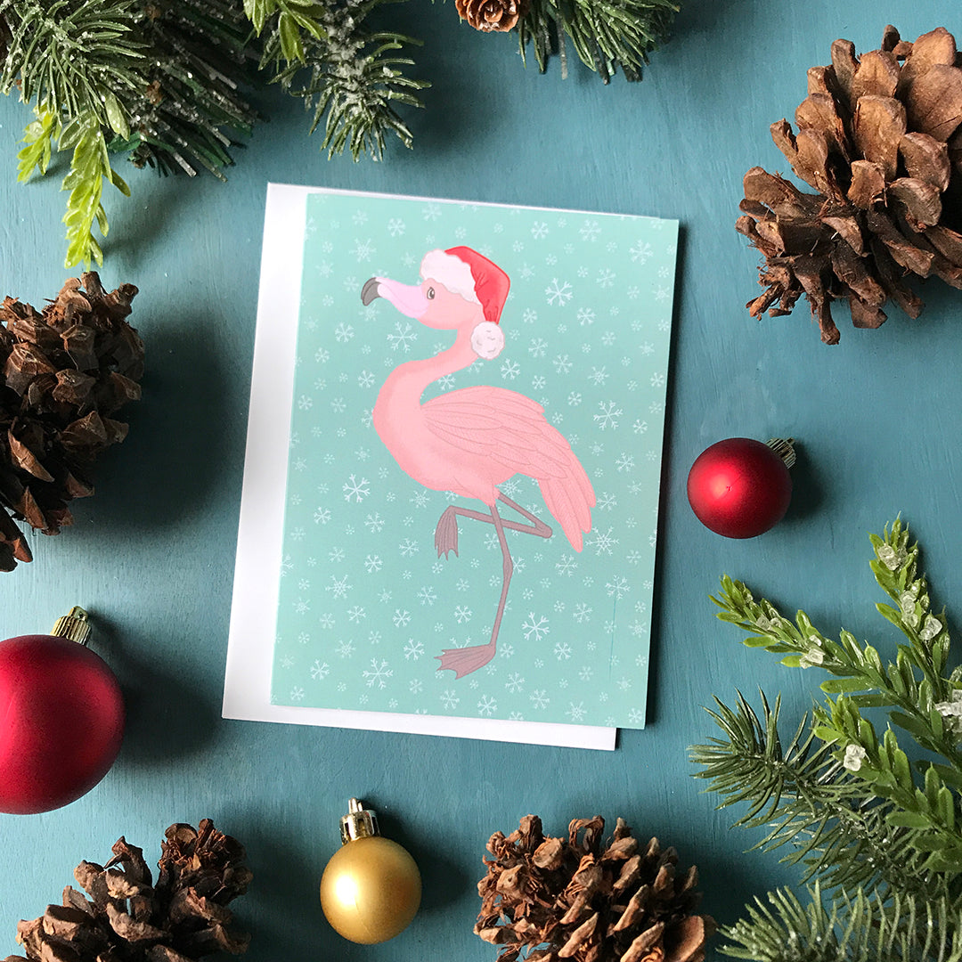 A light blue snowflake-patterned card features a pink flamingo wearing a Santa hat and standing on one leg. The card is surrounded by pinecones, ornaments and faux greenery.