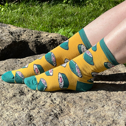 A pair of pale, freckled legs sports a yellow pair of socks patterned with seaweed-wrapped sushi rolls. One foot is outstretched on a rock while the other foot rests on top of that leg, all in front of a grassy background.