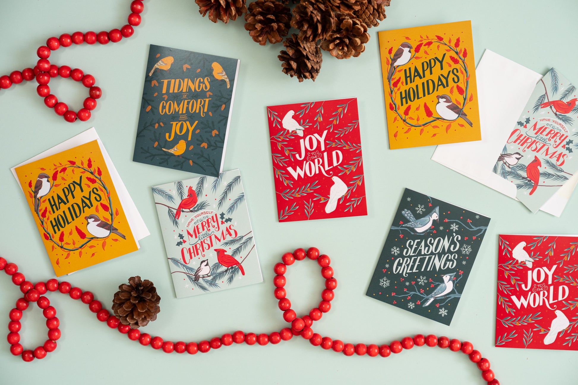 Eight bird-themed Christmas cards appear against a mint background with red beads and pinecones.