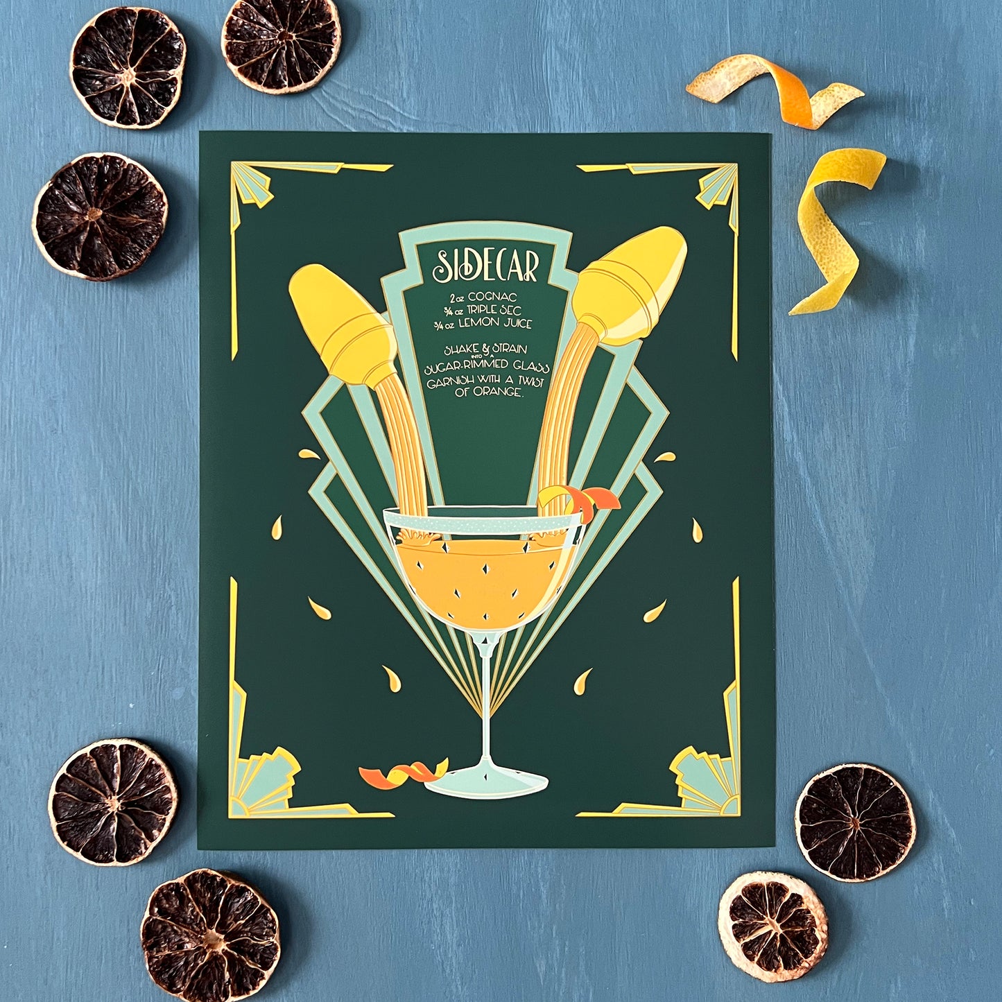 A green and yellow art deco illustration features a hand-lettered recipe for a sidecar as will as images of cocktail shakers and a full glass. The print is lying flat surrounded by dehydrated citrus slices and lemon and orange twists.