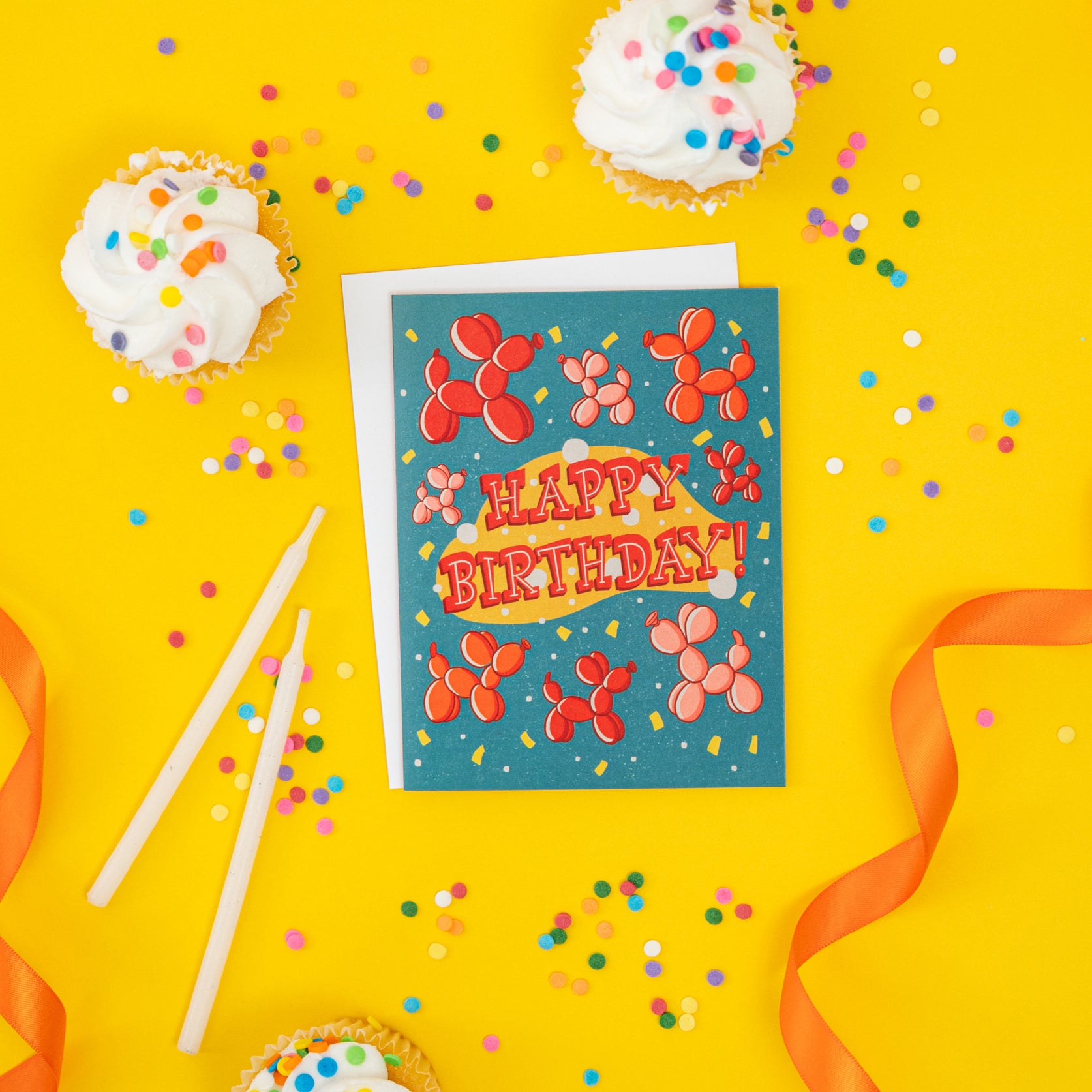 A turquoise birthday card features red, orange and pink balloon dogs, yellow confetti, and the words Happy Birthday! in midcentury-inspired hand lettering. It is on yellow with cupcakes and confetti.