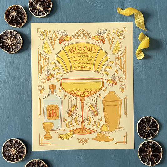 An art-deco inspired print in a yellow and orange colour palette highlights a hand-lettered recipe for a bee's knees cocktail, with a coupe glass, cocktail accessories, lemon twists and bees strewn about the image. The print lies flat and is shown among dehydrated lemon slices and fresh lemon twists.