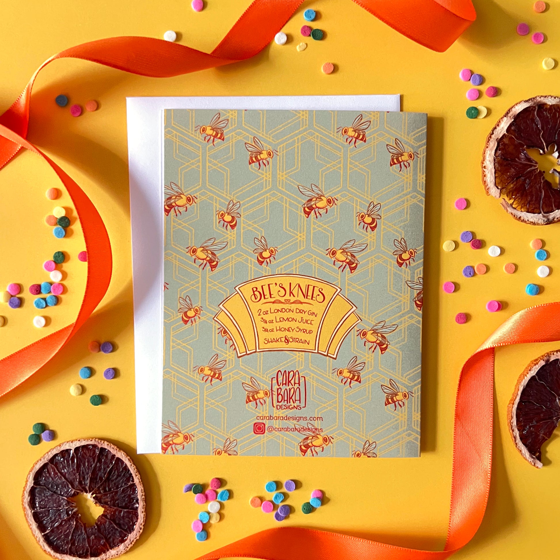 A green greeting card back with art deco hexagons and bees has a hand-lettered cocktail recipe along with Carabara Designs information. The card is on a yellow background with dried citrus, confetti, and orange ribbon.