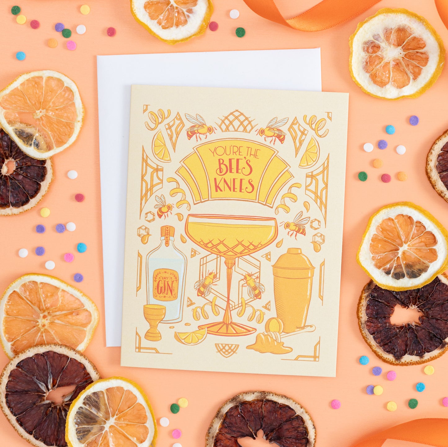 A yellow greeting card with art deco and cocktail details reads You're the Bee's Knees in hand lettering. The card is on an orange background with dried citrus, confetti, and orange ribbon.