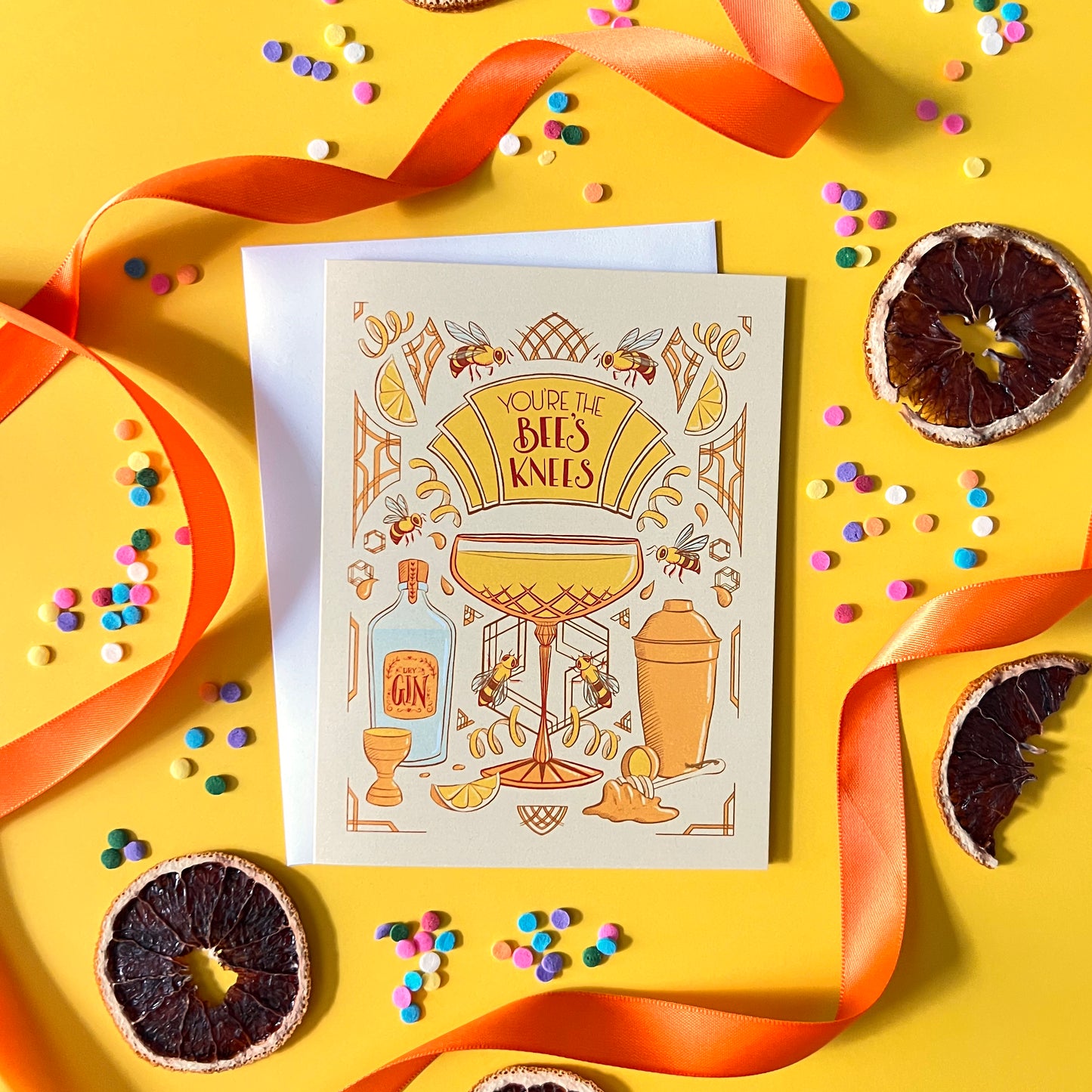 A yellow greeting card with art deco and cocktail details reads You're the Bee's Knees in hand lettering. The card is on a yellow background with dried citrus, confetti, and orange ribbon.