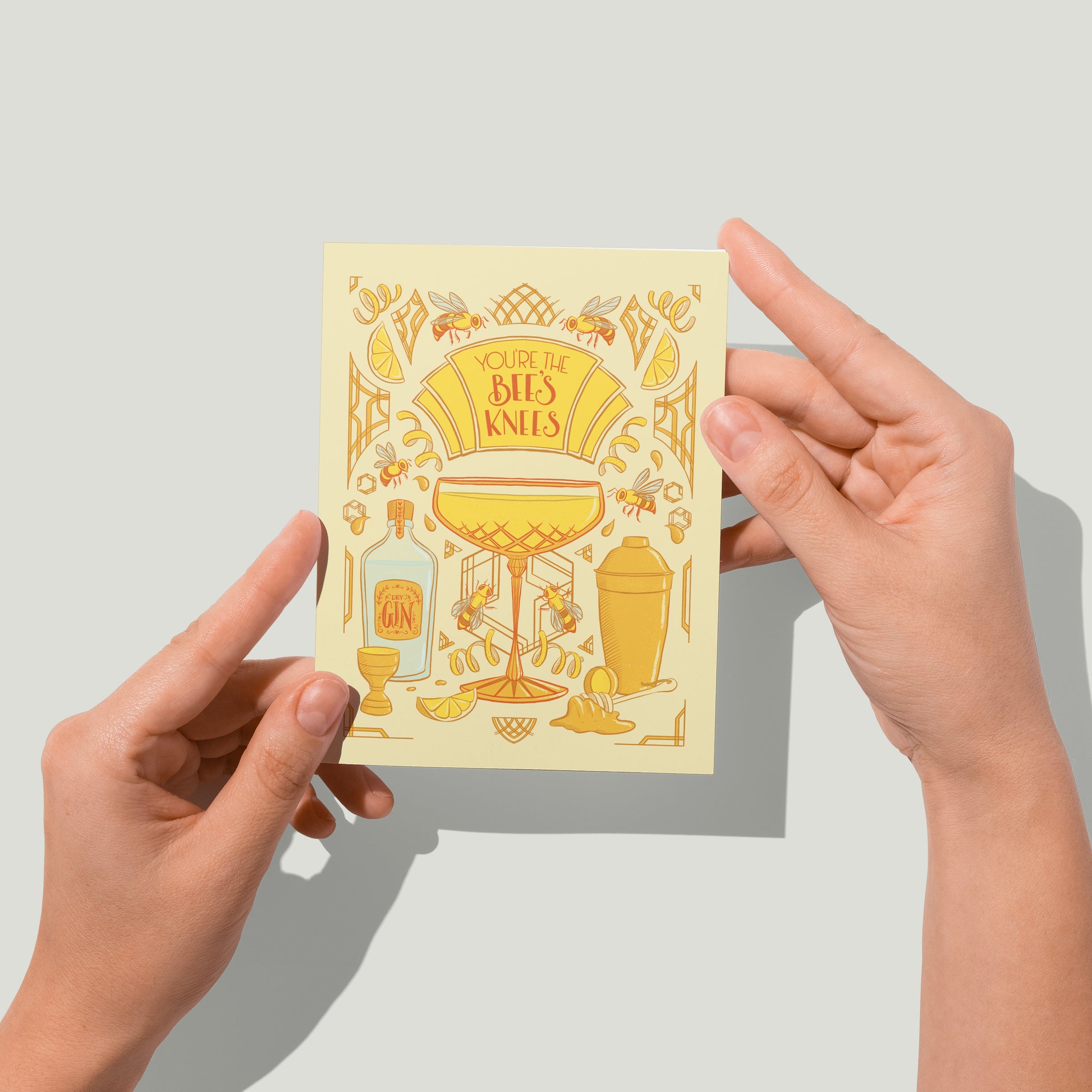 A yellow greeting card with art deco and cocktail details reads You're the Bee's Knees in hand lettering. A pair of hands holds the card up in front of the camera.
