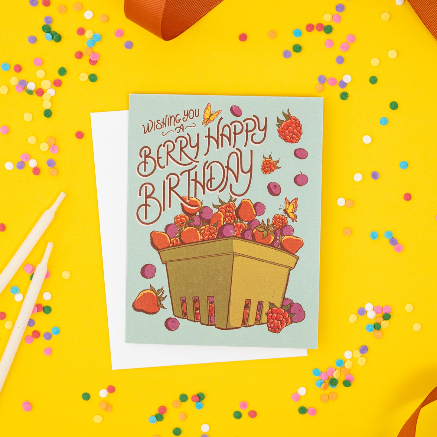 A blue greeting card is covered in strawberries, raspberries and blueberries in a berry basket and reads Have a Berry Happy Birthday. The card is on yellow with confetti, candles and ribbon.