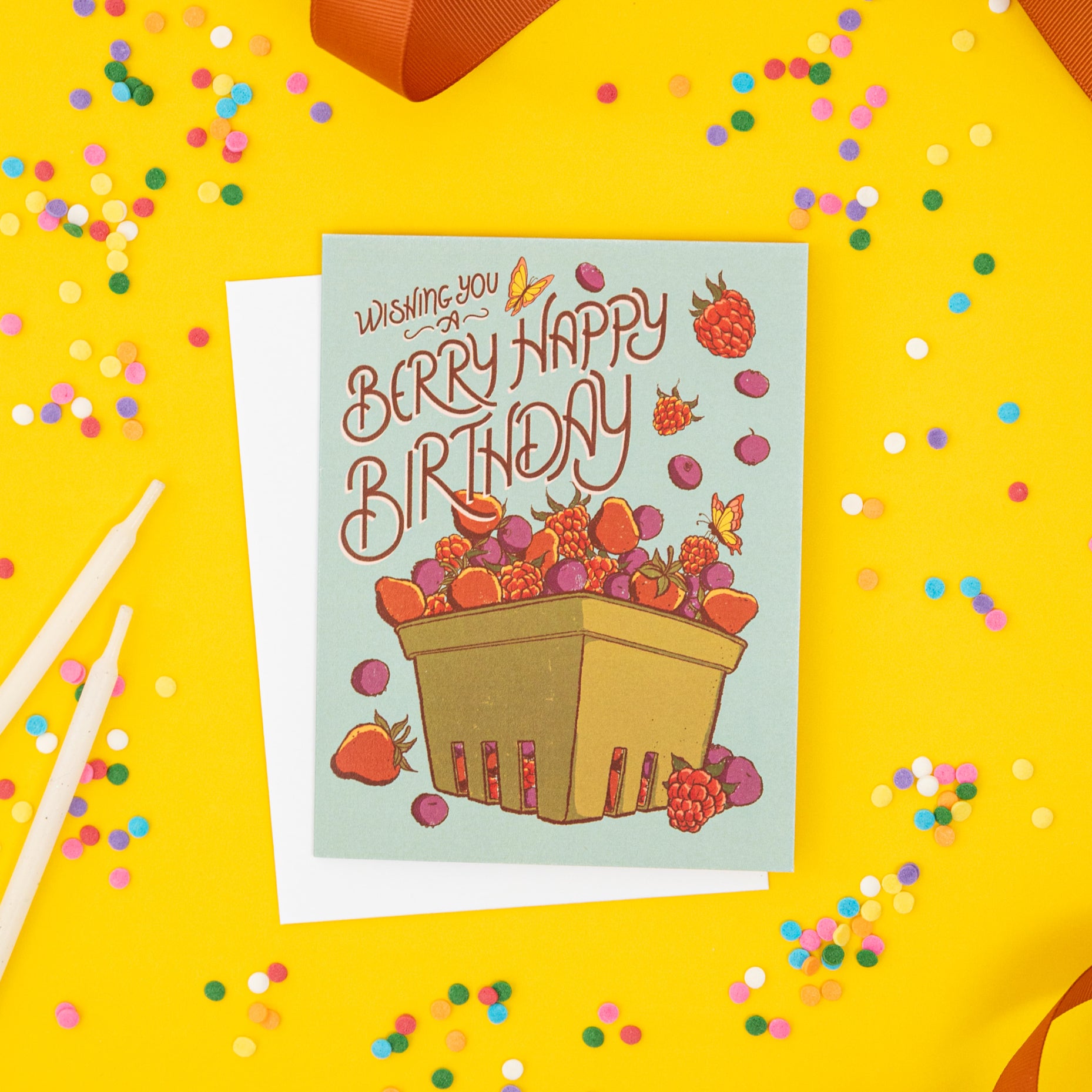 A blue greeting card is covered in strawberries, raspberries and blueberries in a berry basket and reads Have a Berry Happy Birthday. The card is on yellow with confetti, candles and ribbon.