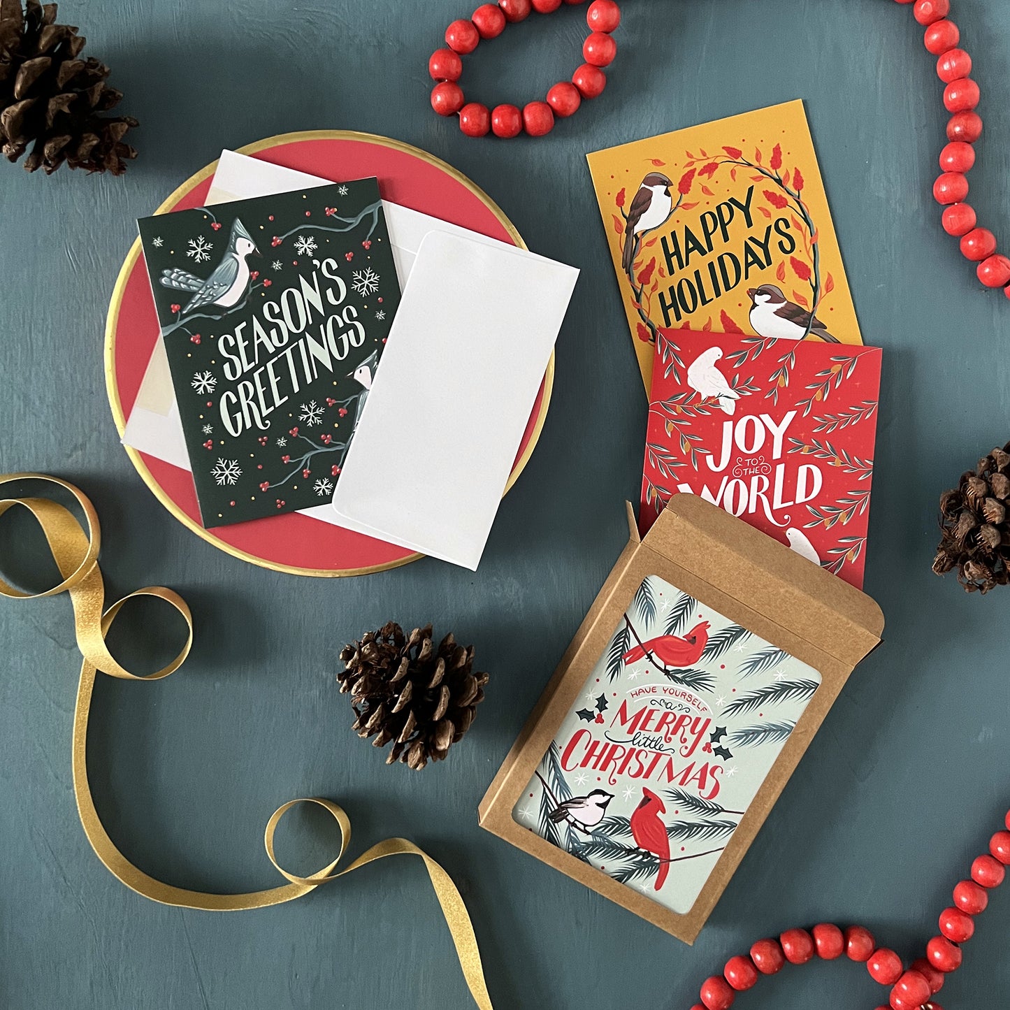 A boxed collection of 4 bird-themed Christmas card designs appears against a festive blue background with pinecones, red beads, and gold ribbon.