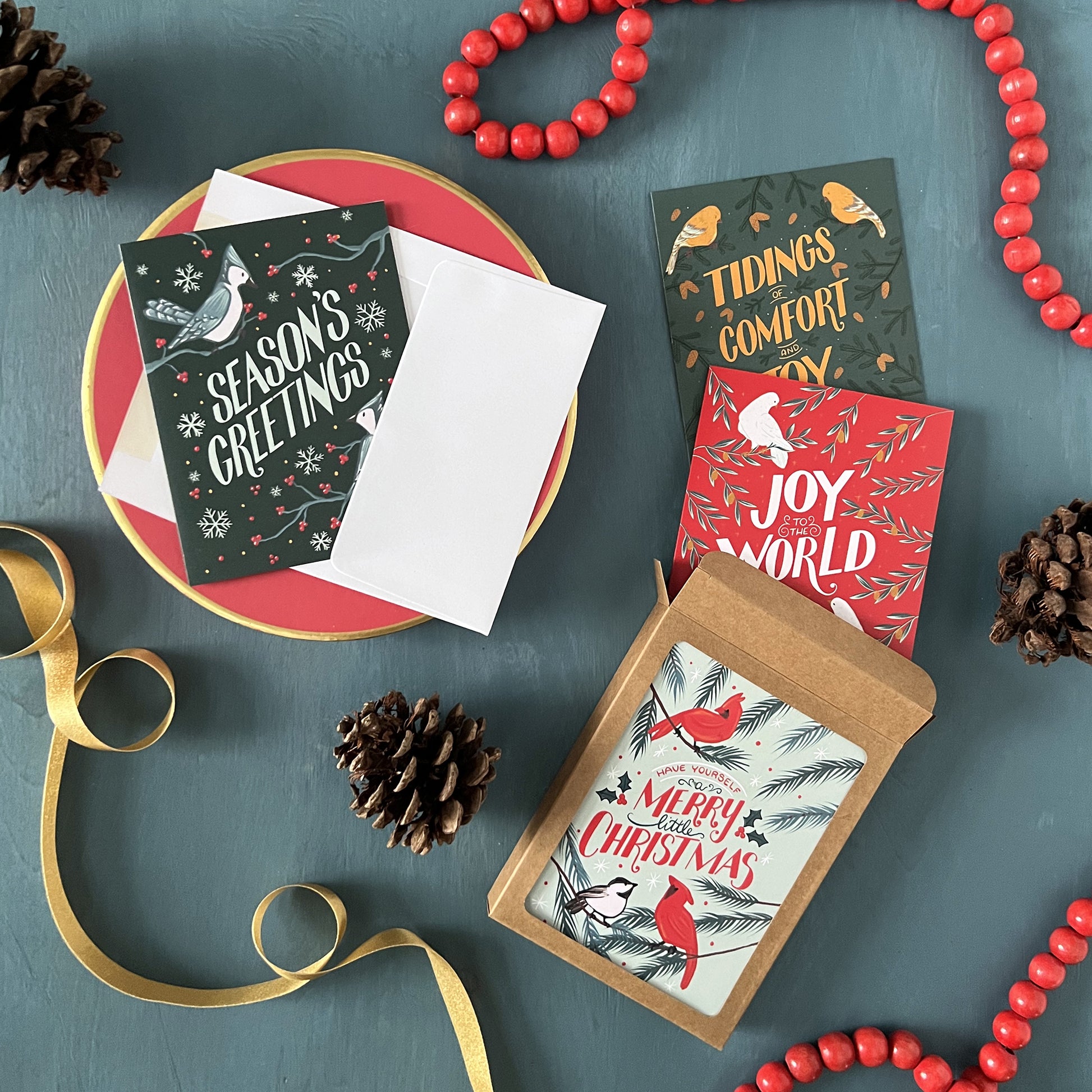 A boxed collection of 4 bird-themed Christmas card designs appears against a festive blue background with pinecones, red beads, and gold ribbon.