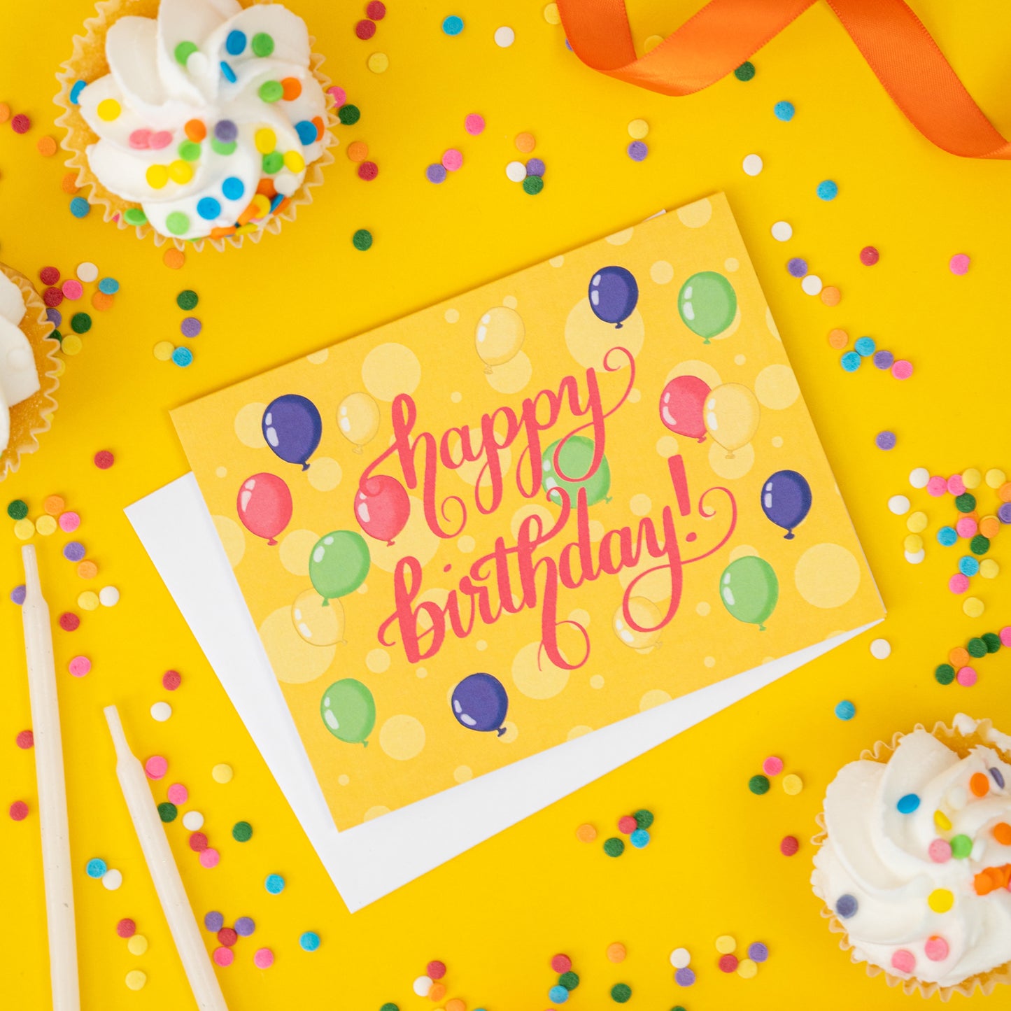 A yellow greeting card is covered with yellow dots and balloons along with the words 'happy birthday' lettered in bright pink. The card sits on yellow with cupcakes and confetti sprinkles.