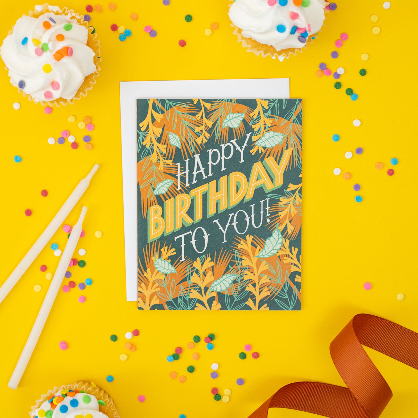 A dark blue greeting card with tropical leaves shows the hand-lettered words 'Happy Birthday to you!'. The card is on yellow with cupcakes and confetti.