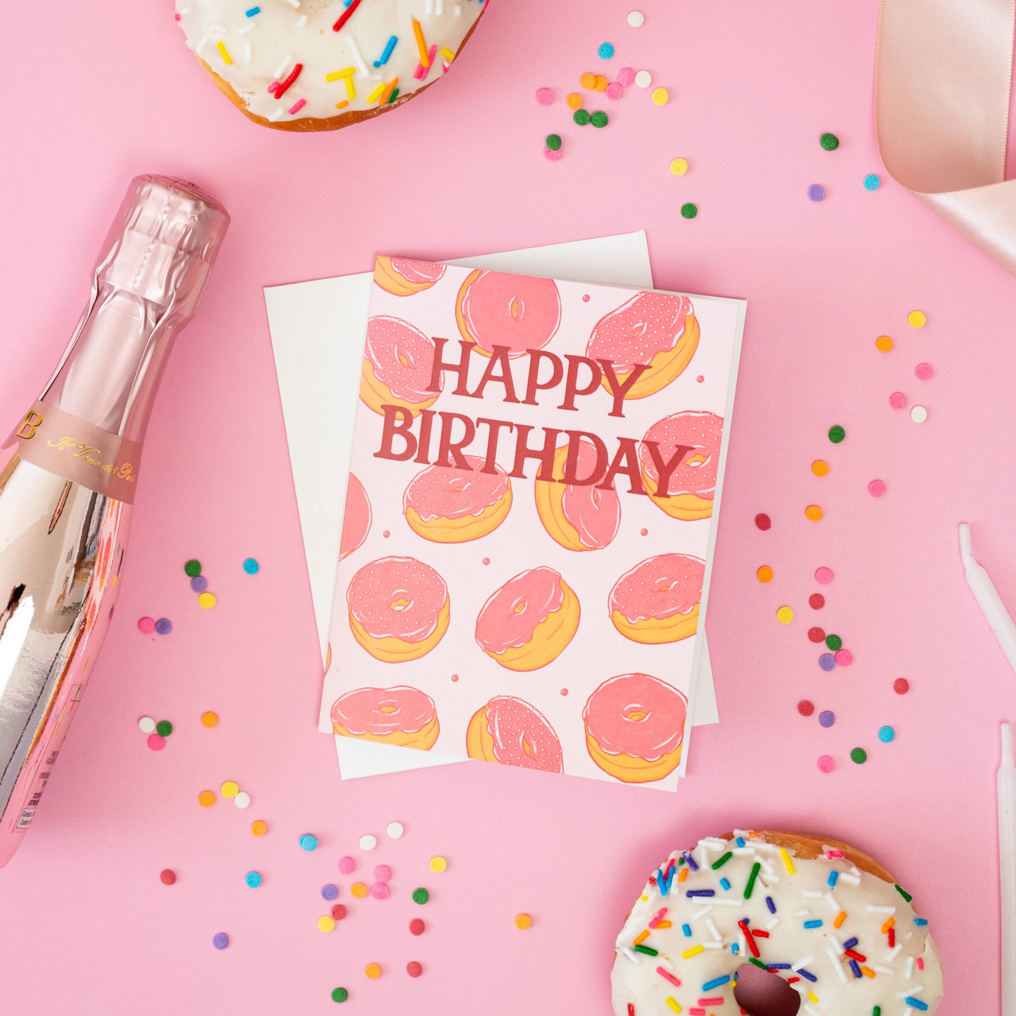 A pink birthday card with doughnuts on it is among rainbow sprinkles, doughnuts and candles on a pink background.