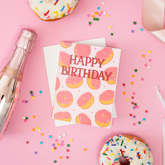 A pink birthday card with doughnuts on it is among rainbow sprinkles, doughnuts and candles on a pink background.
