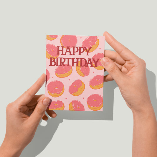 Hands hold a pink birthday card with doughnuts on it up to the camera.