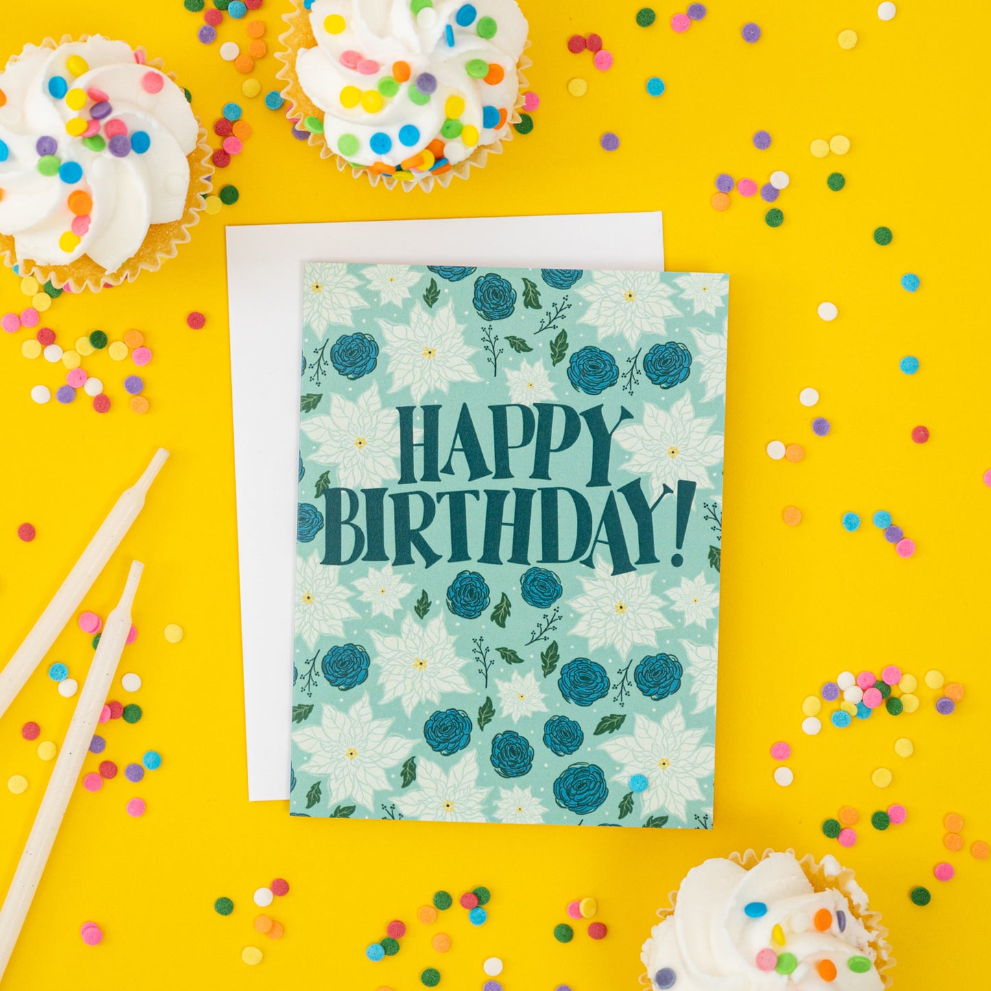 A blue greeting card shows wintry flowers and branches and the words 'happy birthday' in hand lettering. The card is on yellow with confetti, cupcakes and candles.
