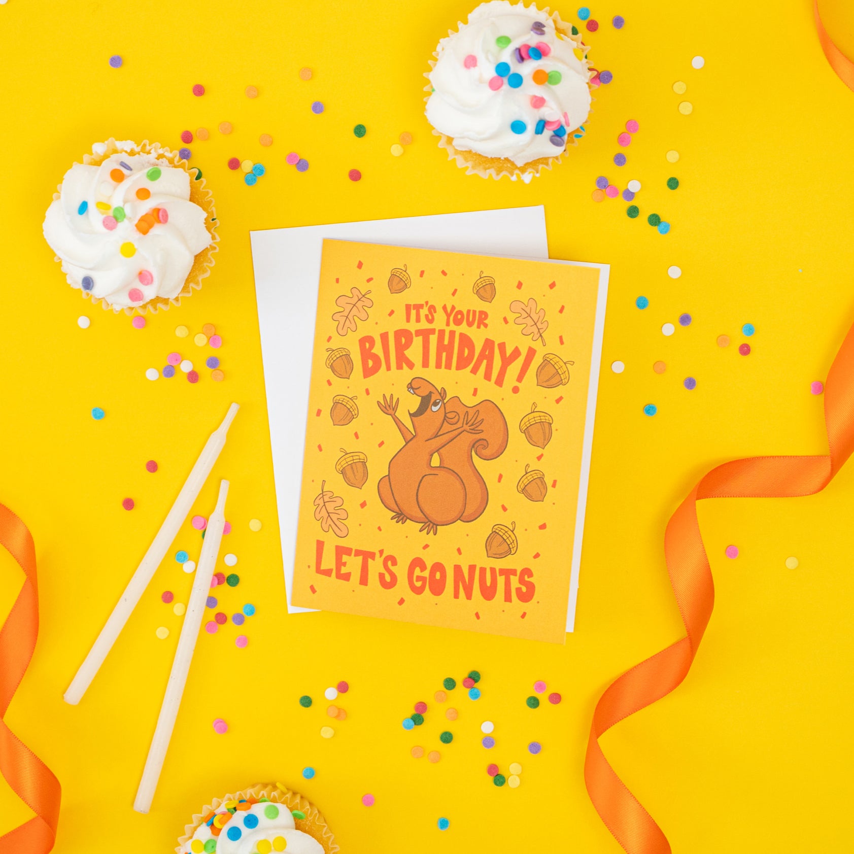 A yellow greeting card shows a joyful squirrel with acorns and confetti along with the words 'it's your birthday! Let's go nuts!'. The card is on yellow and surrounded by confetti, cupcakes and ribbon.