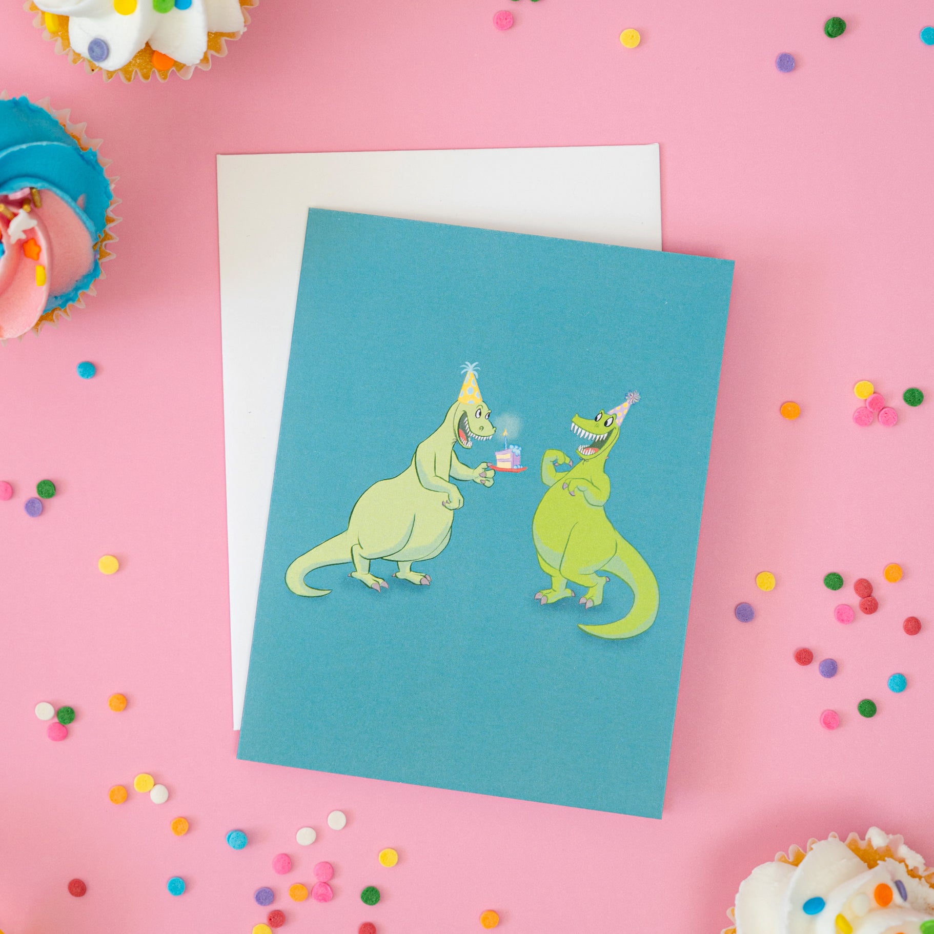 A blue-green greeting card shows an illustration of two grinning green dinosaurs in party hats with cake. The card is on a pink background with cupcakes and confetti sprinkles.