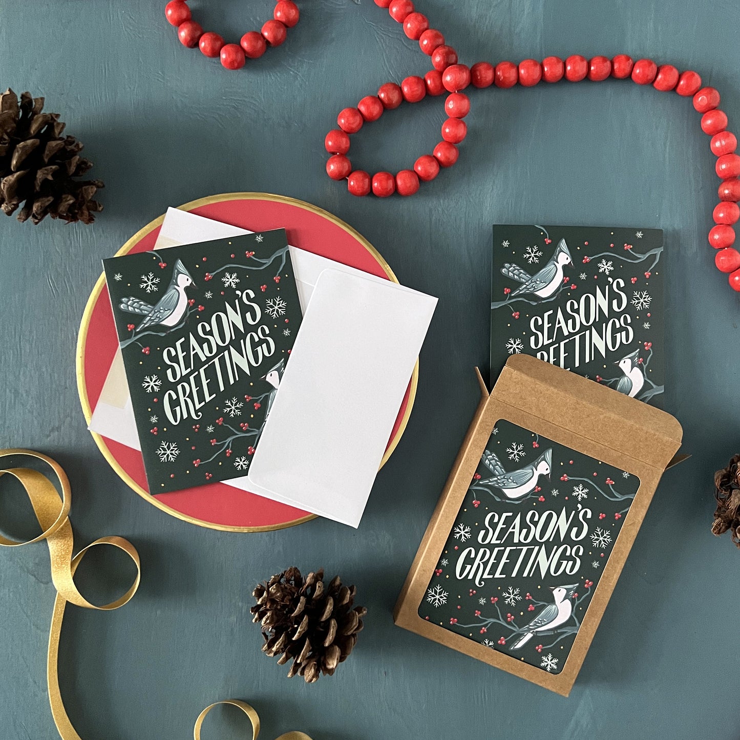 Blue Jay Season's Greetings holiday cards appear on a festive blue background with red beads, pinecones, and ribbon.