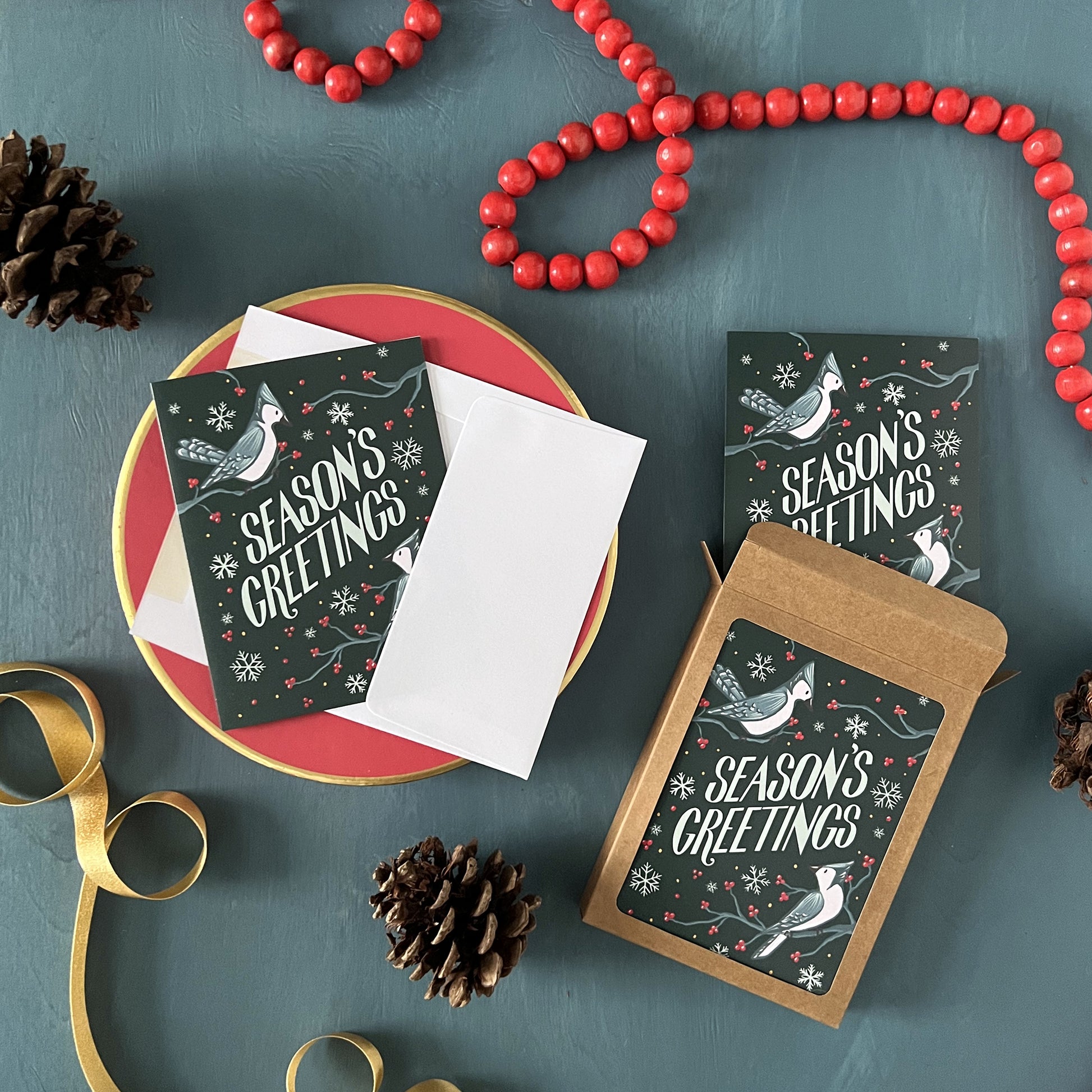 Blue Jay Season's Greetings holiday cards appear on a festive blue background with red beads, pinecones, and ribbon.