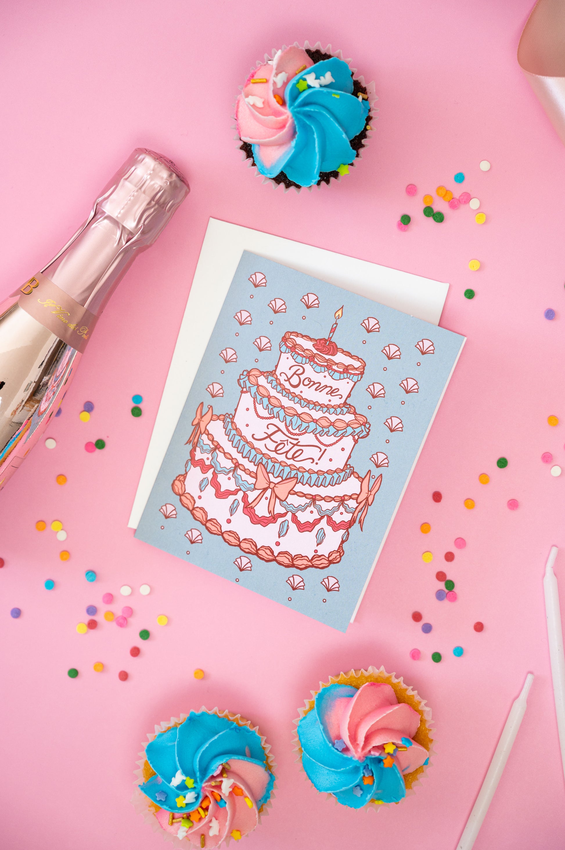 A greeting card shows a rococo birthday cake with the words bonne fête and it sits on a pink background with confetti, candles and cupcakes.