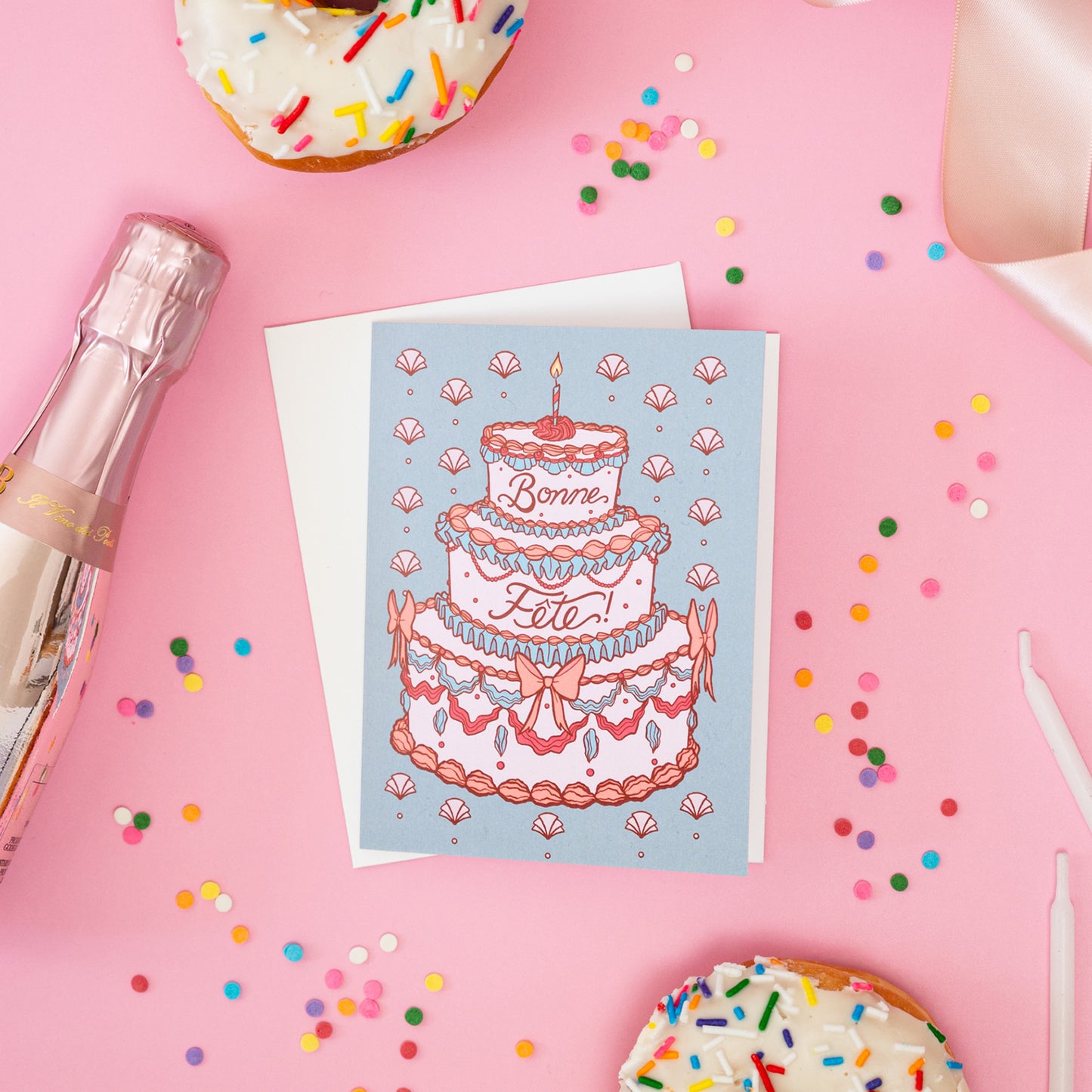 A greeting card shows a rococo birthday cake with the words bonne fête and it sits on a pink background with confetti, candles and doughnuts.