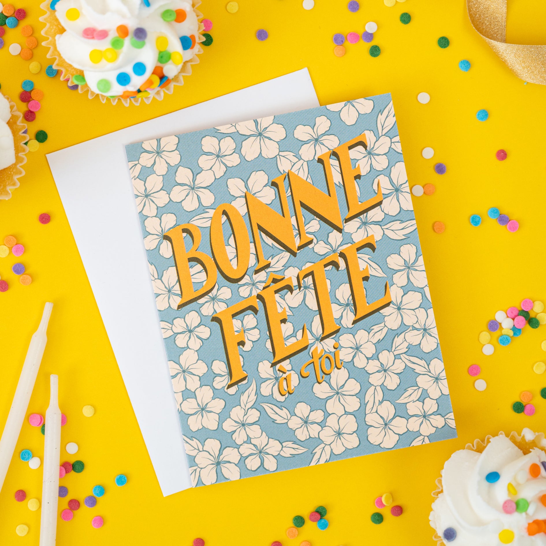 A retro-looking blue greeting card shows creamy flowers and hand lettering that spells out Bonne Fête à toi. It is against a yellow background scattered with confetti sprinkles and cupcakes.