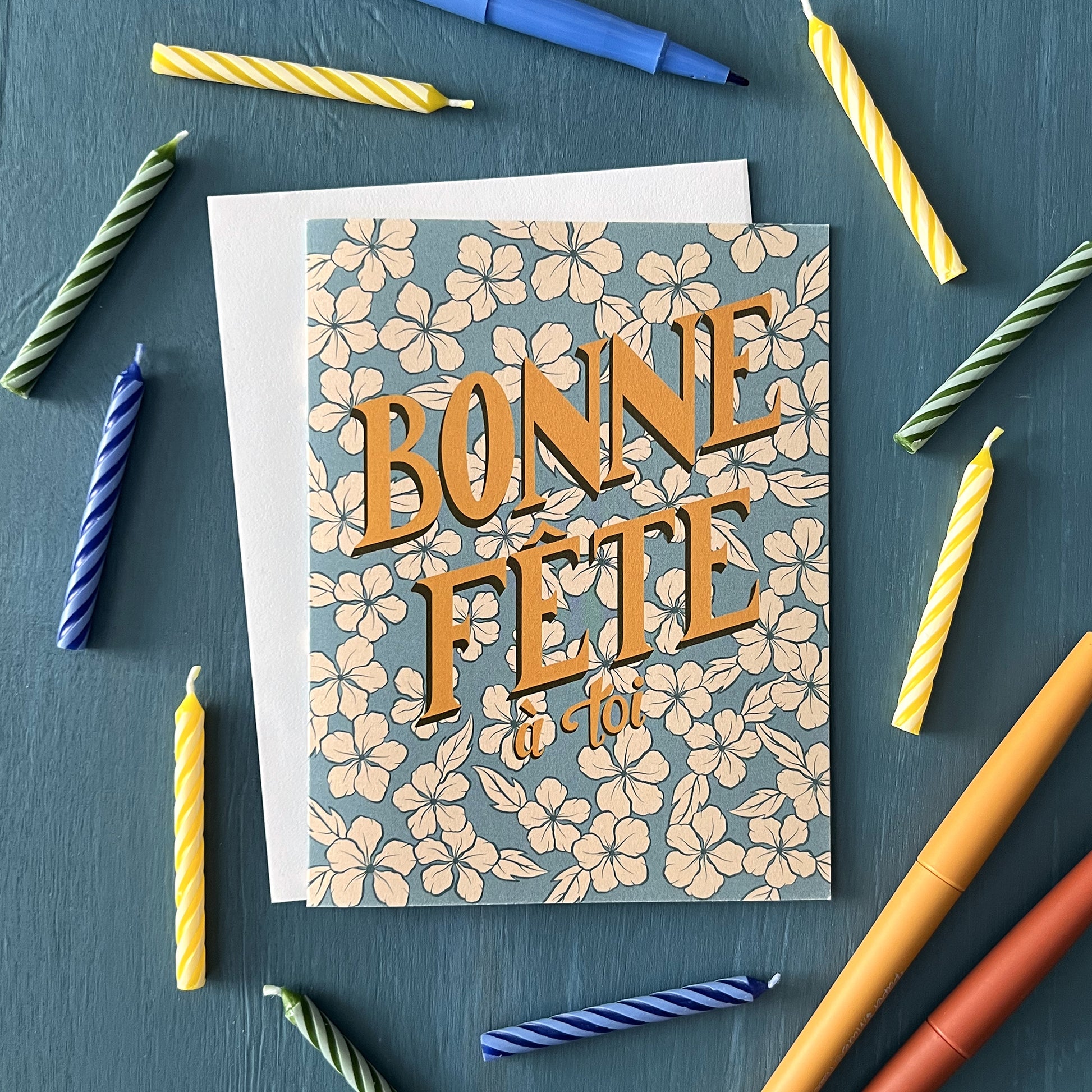 A retro-looking blue greeting card shows creamy flowers and hand lettering that spells out Bonne Fête à toi. The cards is shown against a white envelope and surrounded by birthday candles and pens.