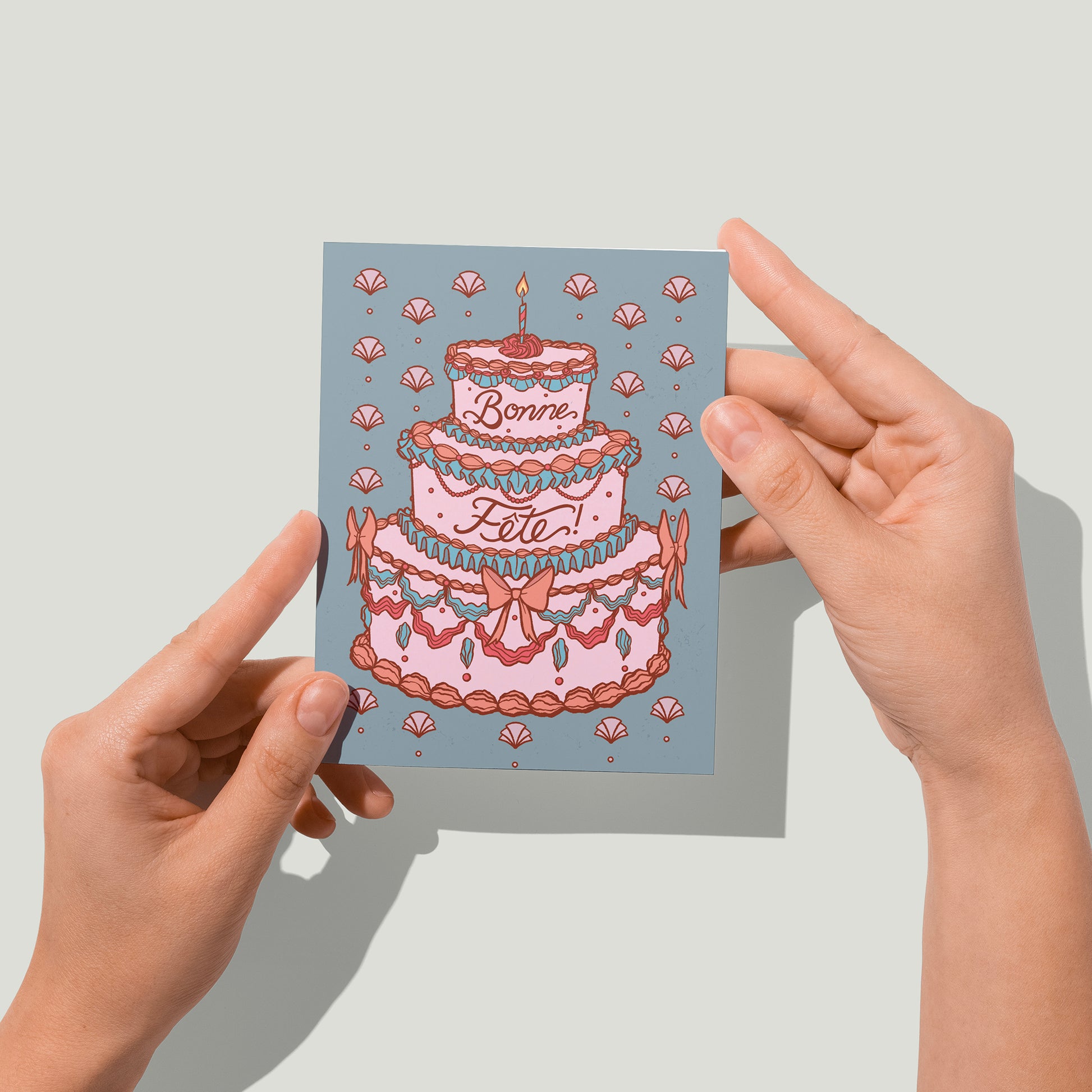 Hands hold a blue birthday card featuring a decadent rococo cake and French lettering up to the camera.