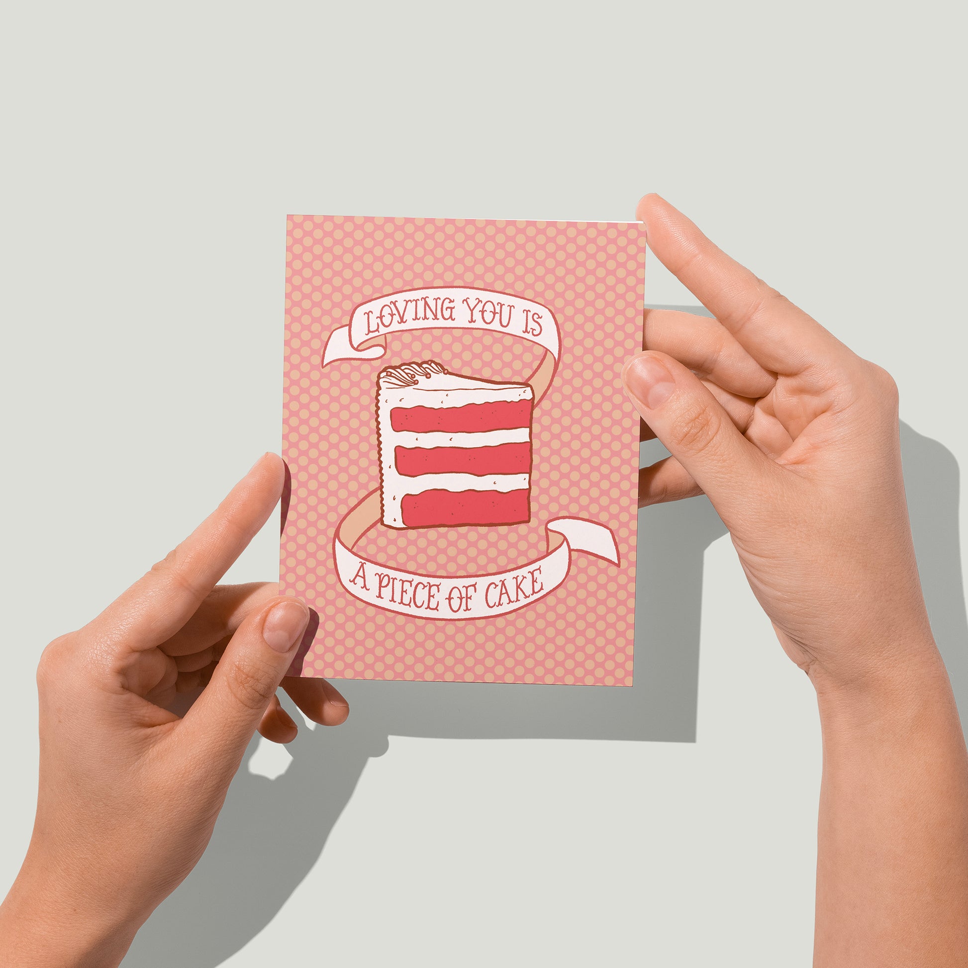 A pink polka dot greeting card shows an illustration of a triple-layer slice of red velvet cake and a banner that reads loving you is a piece of cake in hand lettering. A pair of hands holds the card up in front of the camera.