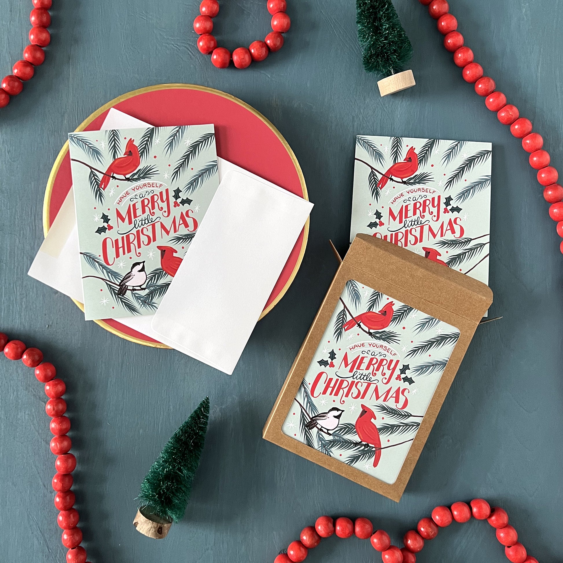 Cardinal and chickadee Christmas greeting cards appear on a festive blue background with red beads and bottle brush trees.