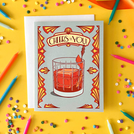 A greeting card says 'cheers to you' in art deco lettering above an old fashioned. The card is on yellow with confetti and candles.