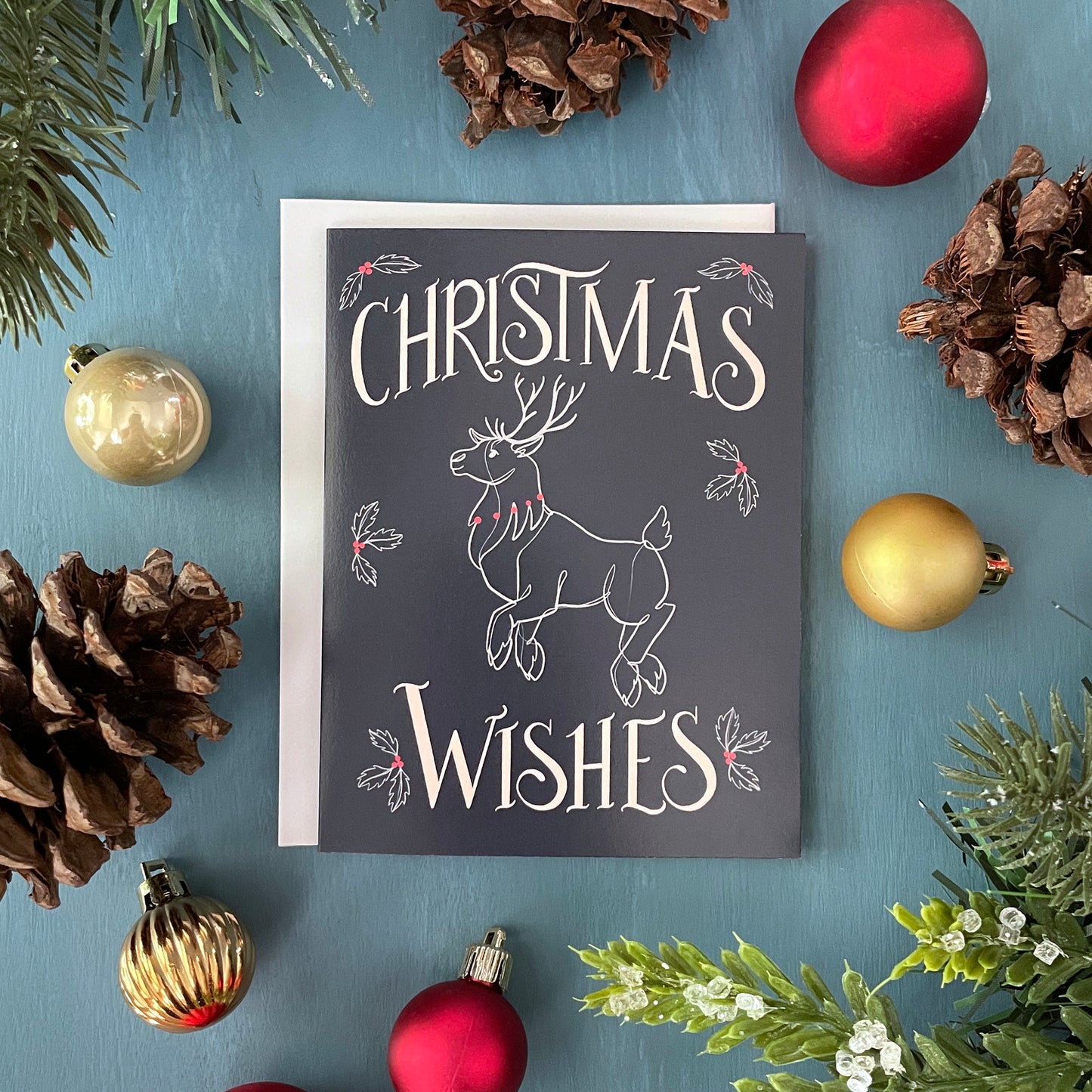 A navy greeting card depicts a flying reindeer in white and red with the word Christmas at the top and Wishes at the bottom. The card is surrounded by faux greenery, pinecones, and Christmas ornaments.