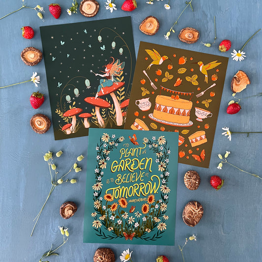 Three cottagecore prints that each include butterflies, leaves, and flowers are on a blue surface surrounded by strawberries, dried mushrooms, and daisies.