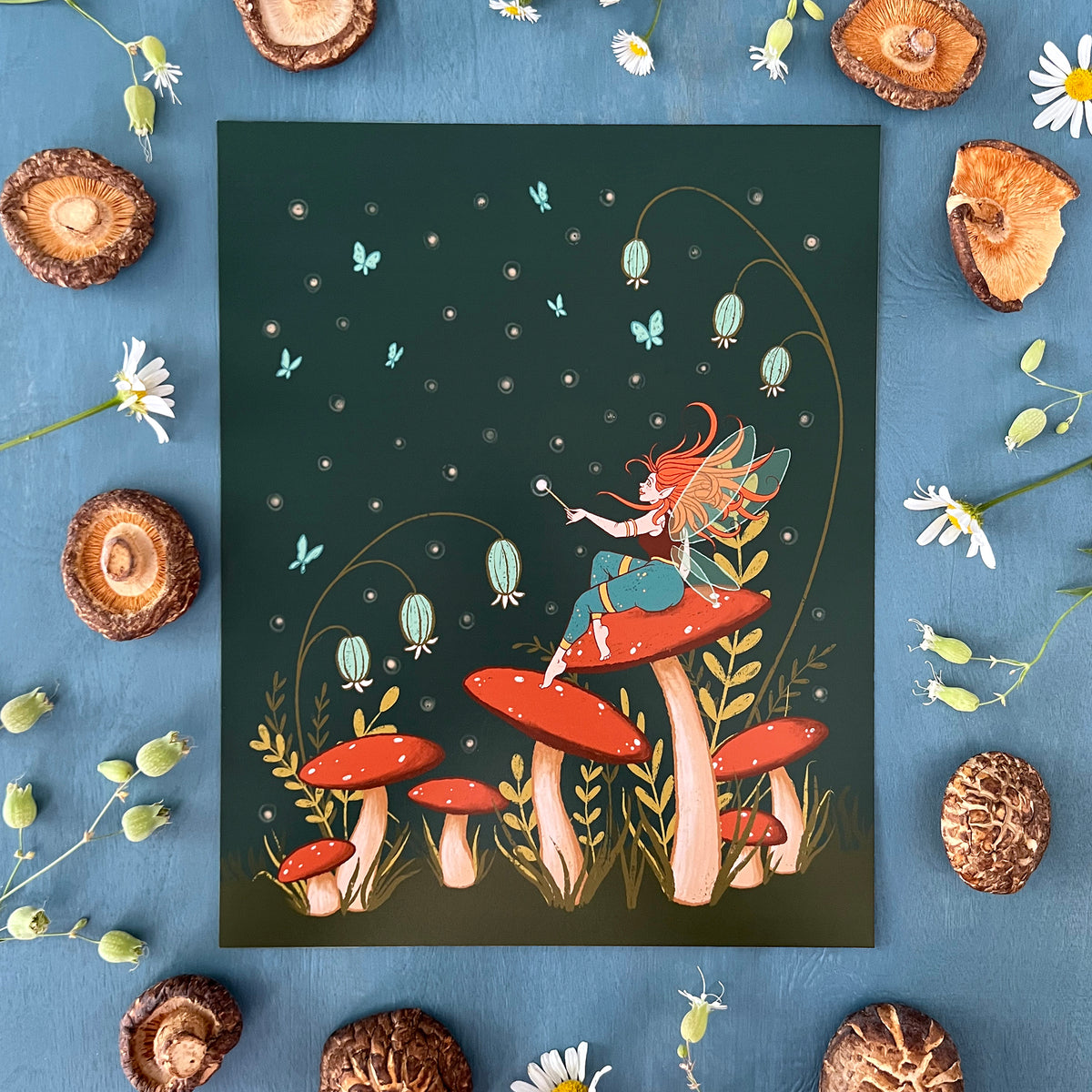 A cottagecore-style illustration shows a winged fairy with turquoise pants sitting on a mushroom in a field of mushrooms, wildflowers and blue butterflies at night. The print is surrounded by wildflowers and mushrooms.