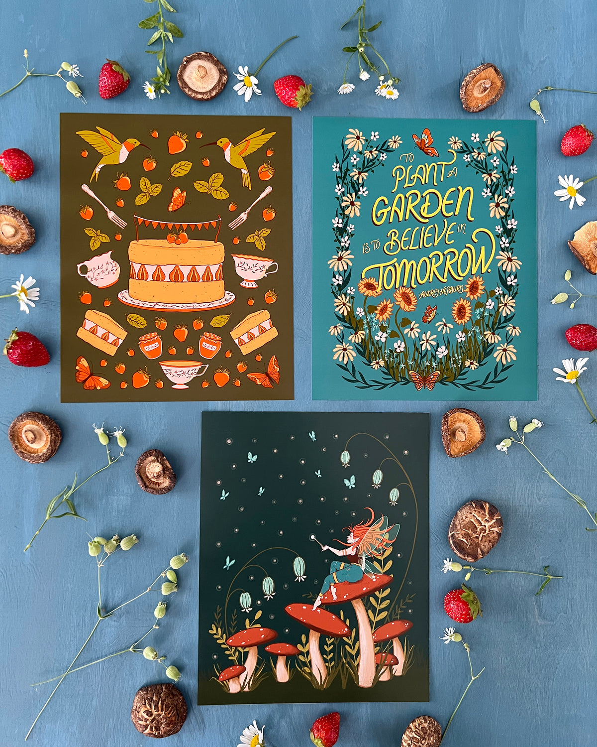 Three cottagecore prints that each include butterflies, leaves, and flowers are laid flat on a blue surface surrounded by strawberries, dried mushrooms, and daisies.