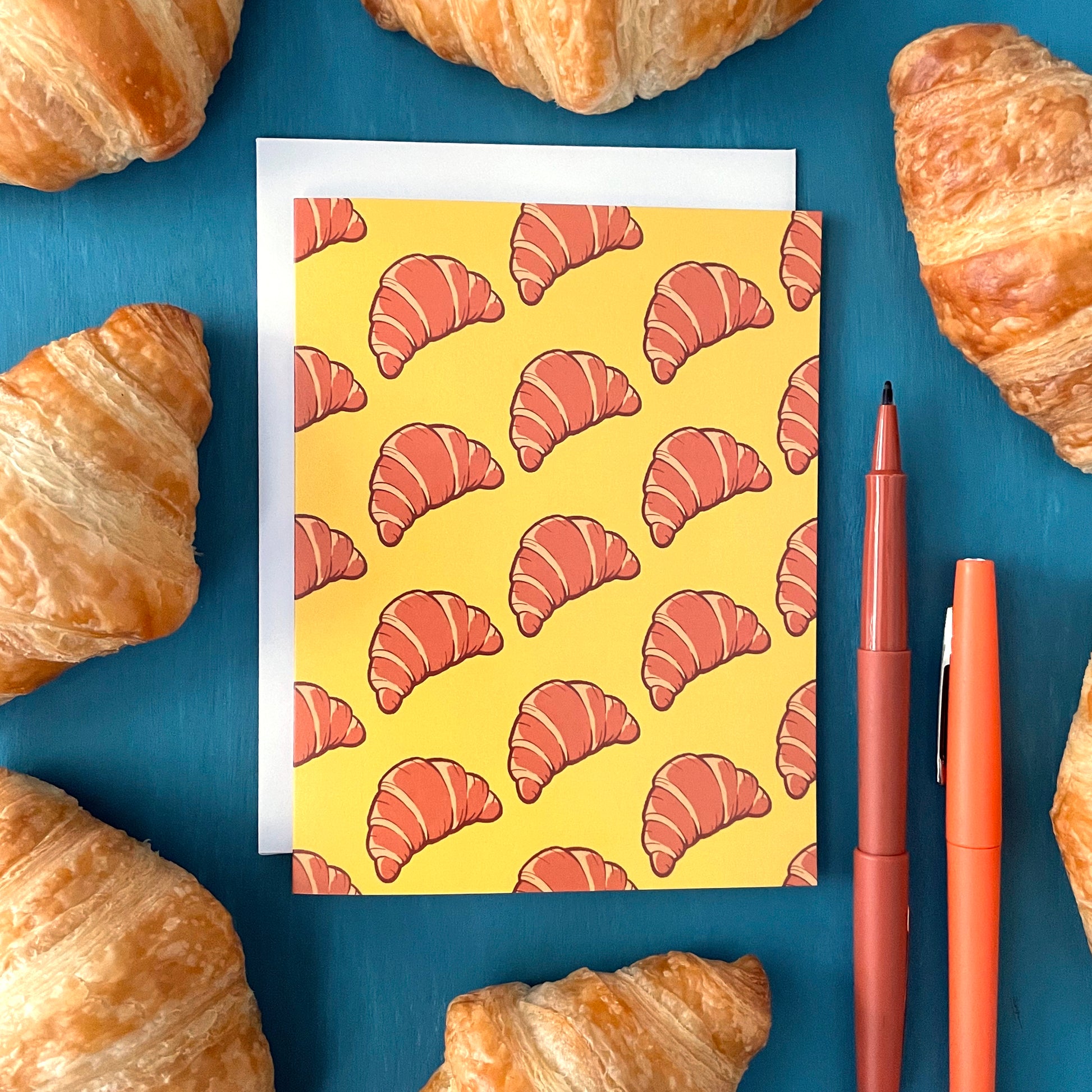 A yellow greeting card is patterned with drawings of croissants in diagonal lines. The card is surrounded by mini croissants and felt-tip pens on a blue background.