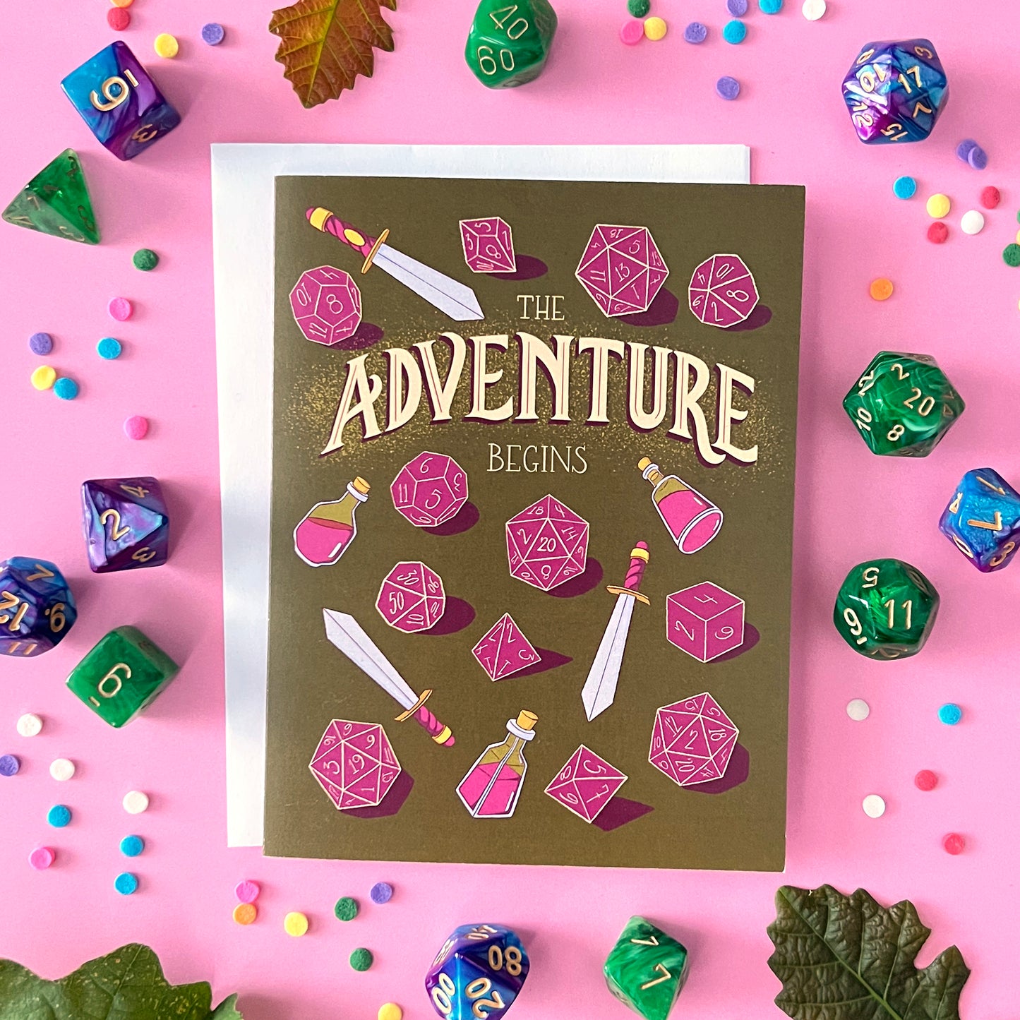A greeting card reads 'the adventure begins' with swords, potions, and dice against pink surrounded by various dice.
