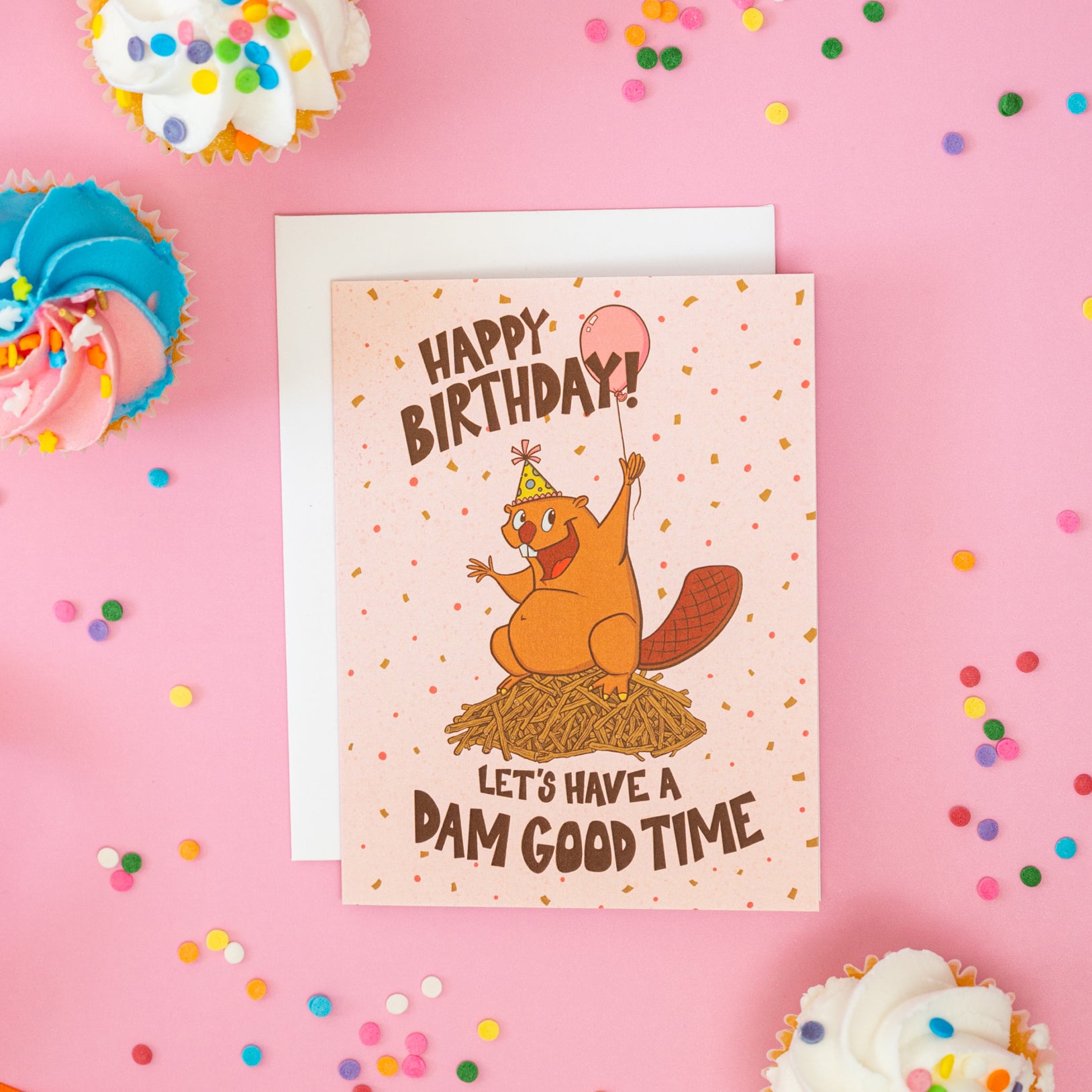 A pink birthday card says Happy Birthday! Let's Have a Dam Good Time in hand-lettering. On the card is a beaver in a party hat on a dam. The card is on pink surrounded by cupcakes and confetti.