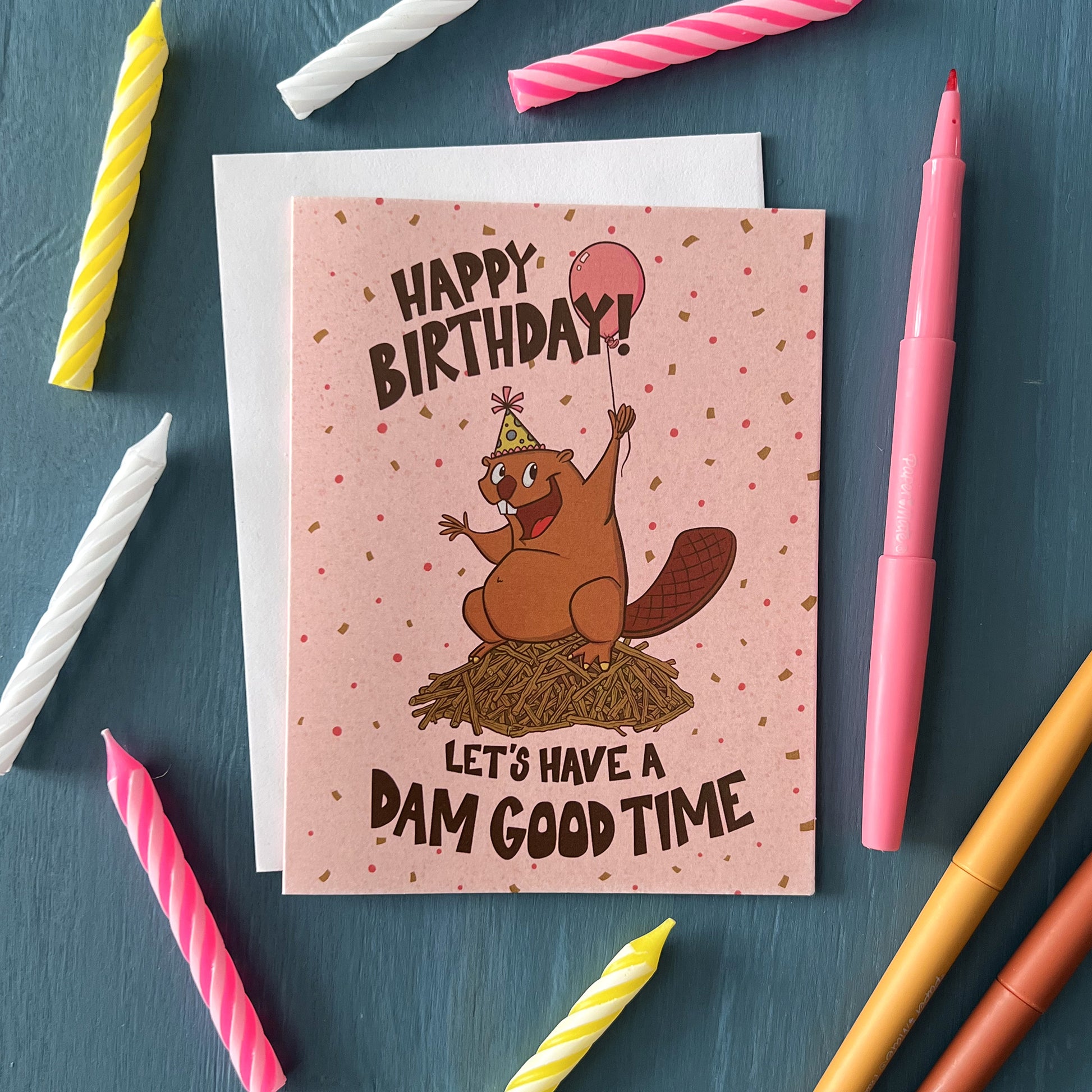 A pink birthday card says Happy Birthday! Let's Have a Dam Good Time in hand-lettering. On the card is a beaver in a party hat on a dam. The card is surrounded by candles and pens.